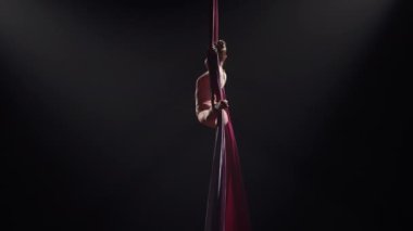 Genç kadın sirk jimnastikçisi havadan ipekle dönüyor ve esneme hareketleri sergiliyor. Arka planda siyah arka planda yükseklikte akrobatik hareketler yapmak zor. Spor, akrobatik, sirk okulu videoları. Yavaş.