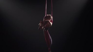 Genç kadın sirk jimnastikçisi havadaki ipeğe baş aşağı asılı duruyor ve hareketin zarafetini gösteriyor. Arka planda siyah bir arka planda yükseklikte akrobatik hareketler yapmak zor. Yavaş çekim.