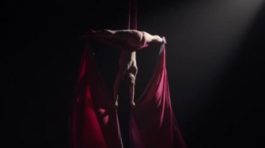 Genç kadın sirk jimnastikçisi havadaki ipeğe baş aşağı asılmış. Akrobat yükseklikte numaralar yapar, kırmızı ipek arka planda siyah arka planda kanatlar gibi çırpınır. Yavaş çekim.