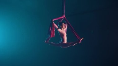 Esnek bir hava jimnastikçisi hava ipeği üzerinde sicim yapar. Genç bir bayan akrobat mavi arka planda siyah bir arka planda dengede duruyor. Spor, akrobatik, sirk okulu videoları. Yavaş çekim.