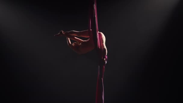 Ung kvinnelig sirkusturner snurrer på luftsilke og viser strekking. Vanskelige akrobatiske stunt i høyden på svart bakgrunn med baklys. Video til sport, akrobatikk, sirkusskole. Langsom. – stockvideo