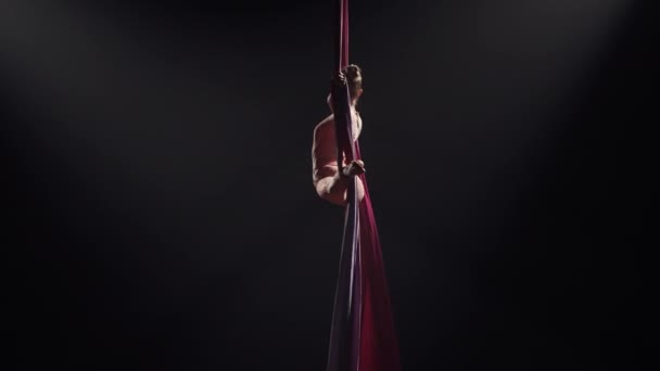 Joven gimnasta de circo femenino gira sobre seda aérea y demuestra estiramiento. Difíciles acrobacias en altura sobre fondo negro con contraluz. Vídeo para deportes, acrobacia, escuela de circo. Despacio.. — Vídeo de stock