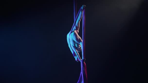 Equilibrio circo gimnasta gira colgando boca abajo en la seda aérea y demuestra estiramiento. Trucos acrobáticos difíciles con lienzo a la altura sobre fondo negro con luz de fondo azul. Movimiento lento. — Vídeos de Stock