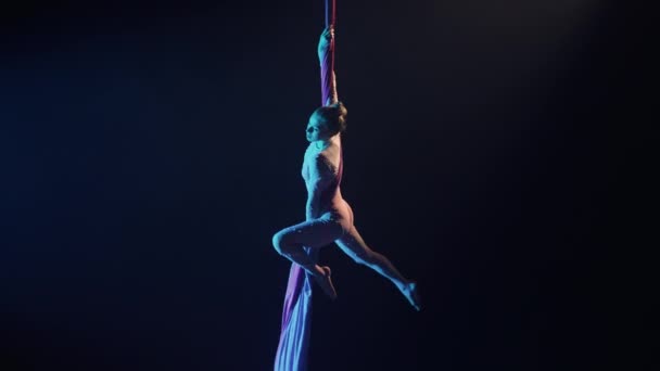 Um talentoso artista de circo feminino equilibra-se em seda arejada. Jovem ginasta aérea mulher executa acrobacias complexas em altura em um fundo estúdio preto com luz de fundo azul. Movimento lento. — Vídeo de Stock