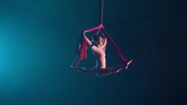 En fleksibel antenne gymnast udfører en sejlgarn på luft silke. En ung akrobat balancerer på en sort baggrund med blåt baggrundslys. Video til sport, akrobatisk, cirkusskole. Langsom bevægelse. – Stock-video