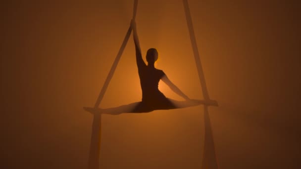 Silhouette af en yndefuld antenne gymnast udfører akrobatiske stunts og sejlgarn på antenne silke. En ung kvinde optræder med cirkusshow i en højde på en røget mørk baggrundsbelyst baggrund. Langsom bevægelse. – Stock-video