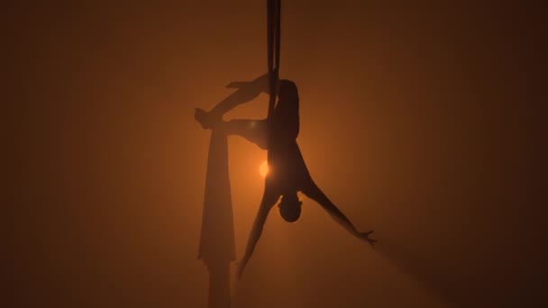 Silhouette af en yndefuld antenne gymnast udfører akrobatiske stunts på antenne silke. En ung kvinde optræder med et cirkusshow i en højde på en røget mørk baggrundsbelyst baggrund. Langsom bevægelse. – Stock-video