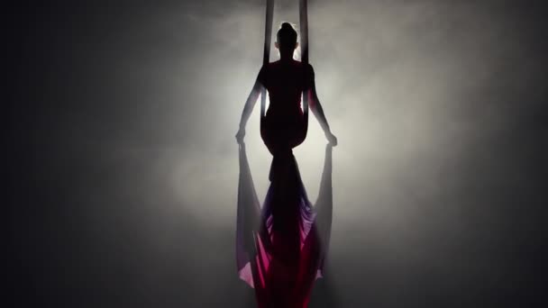 La silueta de gimnasta de circo femenino gira sobre seda aérea. Mujer joven acróbata realiza trucos a una altura, aleteo de seda roja como alas sobre fondo negro ahumado con luz de fondo. Movimiento lento. — Vídeo de stock