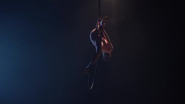 Genç bir kadın jimnastikçi sirk kubbesinin altındaki bir hava çemberinde akrobatik hareketler sergiliyor. Koyu dumanlı bir stüdyoda mavi arka ışıklandırması olan heyecan verici bir akrobatik gösteri. Yavaş çekim.