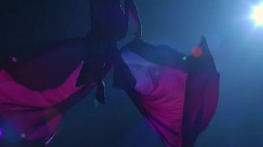 Hava sirki jimnastikçisinin silueti havadan ipekle gösteri yapıyor ve kanat gibi sallanıyor. Genç bayan akrobat tarafından sahnelenen güzel bir sirk gösterisi. Siyah arka planda, mavi arka planda. Yavaş çekim.