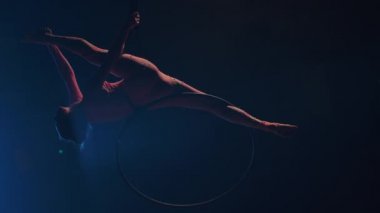 Genç bir kadın jimnastikçi sirkin kubbesinin altındaki bir hava çemberinde akrobatik hareketler sergiliyor ve esneme hareketleri sergiliyor. Koyu dumanlı bir stüdyoda mavi arka ışıklandırması olan heyecan verici bir akrobatik gösteri. Yavaş çekim.