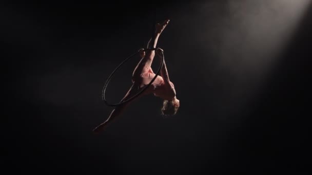 Chica acróbata aérea rotar en el aire en el aro y realiza elementos acrobáticos, demuestra estiramiento y torzal. Equilibrio gimnasta de circo en el fondo negro oscuro estudio con contraluz. Movimiento lento. — Vídeo de stock