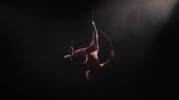 Gadis akrobat udara berputar di udara pada lingkaran dan melakukan elemen akrobatik, menunjukkan peregangan dan benang. Equilibrium sirkus pesenam di pada latar belakang hitam studio gelap dengan cahaya latar belakang. Gerakan lambat. — Stok Video