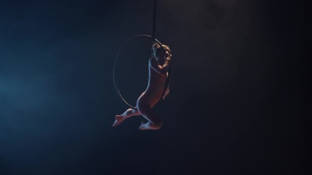 En ung kvinnlig gymnast utför akrobatiska stunts på en luftring under kupolen på en cirkus. Spännande akrobatisk show i en mörk rökig studio med blått bakgrundsljus. Långsamma rörelser. — Stockvideo