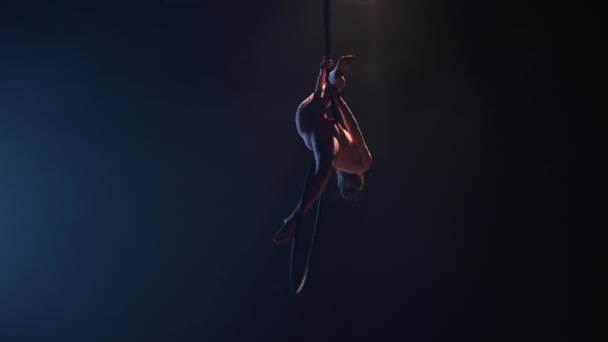 Seorang pesenam wanita muda melakukan aksi akrobatik di atas ring udara di bawah kubah sirkus. Pertunjukan akrobatik yang menarik di studio gelap berasap dengan latar belakang biru. Gerakan lambat. — Stok Video