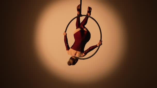 Um belo aerialista está girando de cabeça para baixo em um anel aéreo alto sob a cúpula do circo. Uma jovem realiza acrobacias acrobáticas em um fundo de estúdio escuro retroiluminado. Movimento lento. — Vídeo de Stock