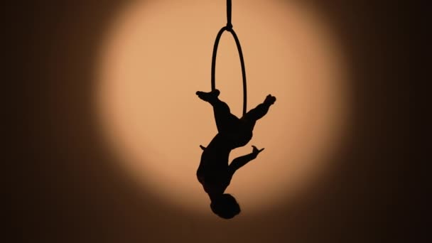 Den sorte silhuet af en antenne gymnast udfører akrobatiske tricks og spinding i luften på ringen. En ung kvinde akrobat udfører på mørk studie baggrund med baggrundslys. Langsom bevægelse. – Stock-video