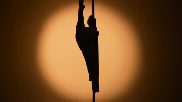 Silhouet van een jonge vrouw voert acrobatische elementen op luchtige zijde. Een luchtturner demonstreert stretching tijdens het uitvoeren van een truc op hoogte in een donkere verlichte studio. Langzame beweging. — Stockvideo