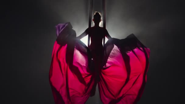 Silhouette of an aerial circus gymnast performing on aerial silk and waving canvas like wings. Beautiful circus show performed by young woman acrobat on black background with backlight. Slow motion. — Stock Video