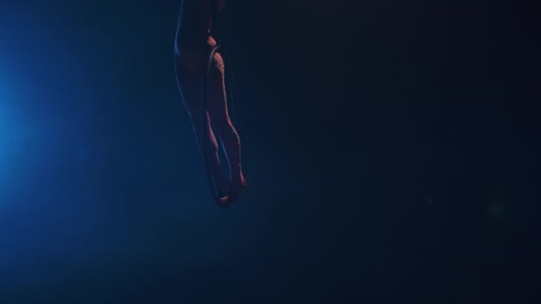 Uma jovem ginasta realiza acrobacias acrobáticas em um arco de ar sob a cúpula de um circo. Excitante show acrobático em um estúdio fumegante escuro com luz de fundo azul. Movimento lento. — Vídeo de Stock