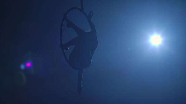 Silhouette of flexible aerial gymnast rotates on an aerial ring and performs acrobatic tricks in the air. Young woman performs with circus show in dark smoky studio with blue backlight. Slow motion. — Stock Video