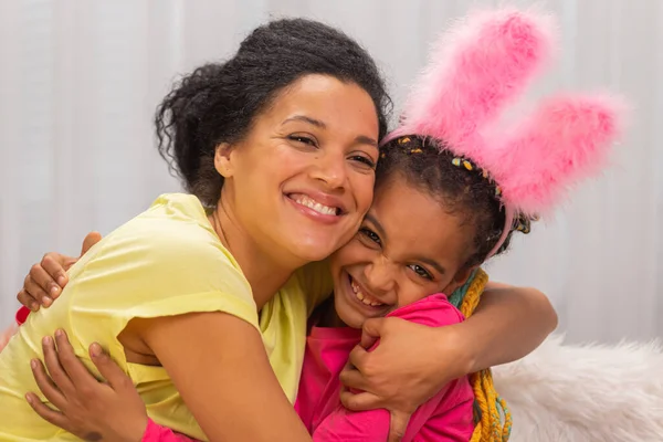 Mamma e figlia con divertenti orecchie da coniglio si abbracciano e sorridono felicemente. Donna afroamericana e bambina sono seduti a tavola in una stanza decorata a festa a casa. Da vicino.. — Foto Stock