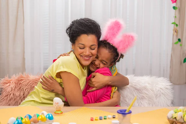 Mamma e figlia con divertenti orecchie da coniglio si abbracciano e sorridono felicemente. Donna afroamericana e bambina sono seduti a tavola in una stanza decorata a festa a casa. Da vicino.. — Foto Stock