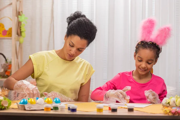 Mamma e figlia dipingono uova usando vernice gialla. Donna afroamericana e bambina sono seduti in camera decorata a casa. La famiglia si sta preparando per le vacanze di primavera. Buona Pasqua. Da vicino.. — Foto Stock