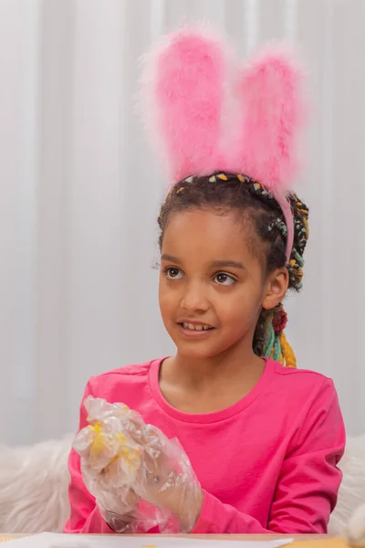 La bambina carina in guanti protettivi trasparenti dipinge un uovo di Pasqua giallo con vernice. Bambino afroamericano sono seduti in camera decorata a casa. Buona Pasqua. Da vicino.. — Foto Stock