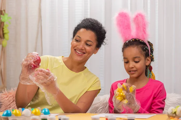 Mamma e figlia in guanti protettivi trasparenti dipingono un uovo di Pasqua rosa e giallo con vernice. Donna afroamericana e bambina sono seduti in camera decorata a casa. Buona Pasqua. Da vicino.. — Foto Stock