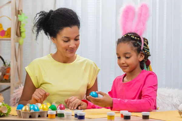 Mamma e figlia con divertenti orecchie di coniglio incollare immagini su uova colorate. Donna afroamericana e bambina sono seduti a tavola in una stanza decorata a festa a casa. Buona Pasqua. Da vicino.. — Foto Stock
