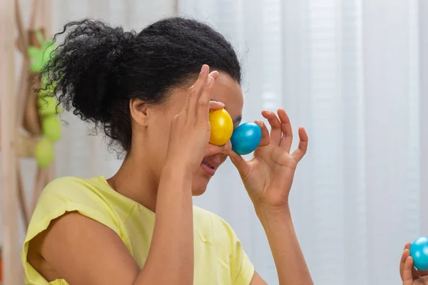 Mamma e figlia con divertenti orecchie da coniglio ridono e si divertono con uova colorate. Donna afroamericana e bambina seduta a tavola in camera decorata a casa. Buona Pasqua. Da vicino.. — Foto Stock