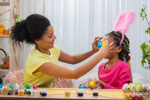 Mamma e figlia con divertenti orecchie da coniglio ridono e si divertono con uova colorate. Donna afroamericana e bambina seduta a tavola in camera decorata a casa. Buona Pasqua. Da vicino.. — Foto Stock