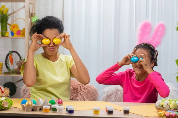 Mamma e figlia con divertenti orecchie da coniglio ridono e si divertono con uova colorate. Donna afroamericana e bambina seduta a tavola in camera decorata a casa. Buona Pasqua. Da vicino.. — Foto Stock