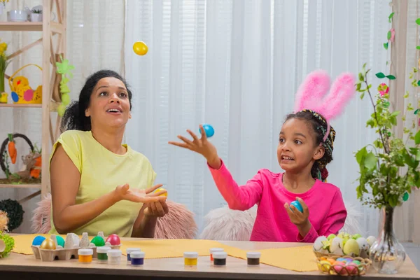 Mamma e figlia con divertenti orecchie da coniglio ridono e si divertono con uova colorate. Donna afroamericana e bambina seduta a tavola in camera decorata a casa. Buona Pasqua. Da vicino.. — Foto Stock