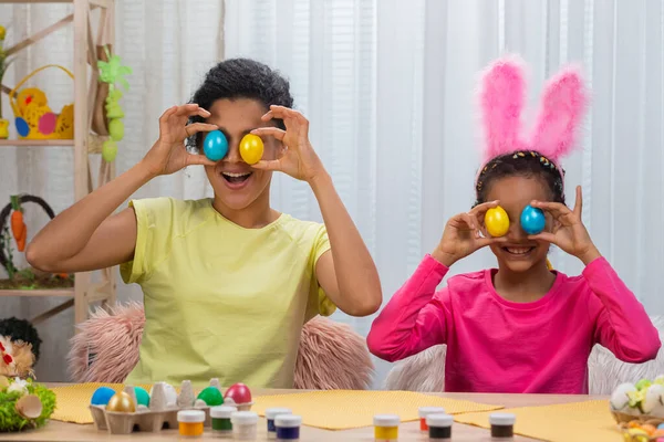 Mamma e figlia con divertenti orecchie da coniglio ridono e si divertono con uova colorate. Donna afroamericana e bambina seduta a tavola in camera decorata a casa. Buona Pasqua. Da vicino.. — Foto Stock