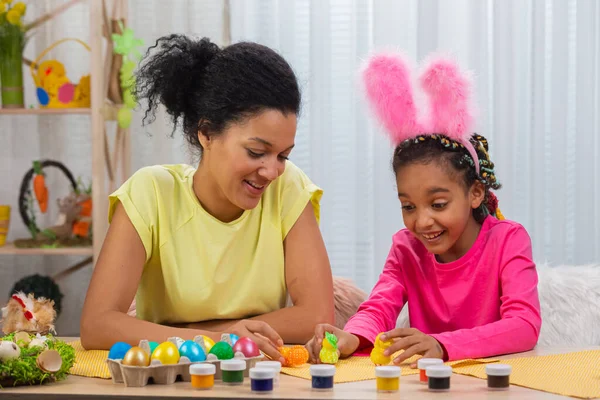 Mamma e figlia con divertenti orecchie di coniglio si divertono con pulcini decorativi. Donna afroamericana e bambina sono seduti a tavola in una stanza decorata a festa a casa. Buona Pasqua. Da vicino.. — Foto Stock