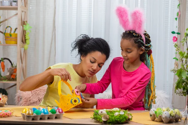 Mamma e figlia con divertenti orecchie da coniglio decorano con fiori cesto di uova. Donna afroamericana e bambina seduta a tavola in una stanza decorata a festa a casa. Buona Pasqua. Da vicino.. — Foto Stock