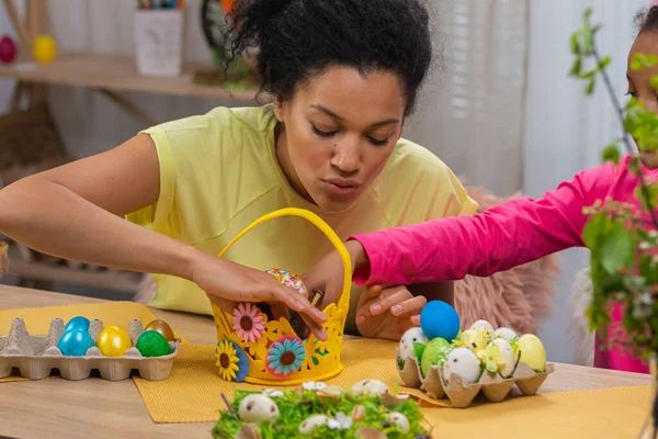 Mamma e figlia mettere torta di Pasqua e uova colorate nel cestino. Donna afroamericana e bambina sedute in camera decorata a casa. Buona Pasqua. Da vicino.. — Foto Stock