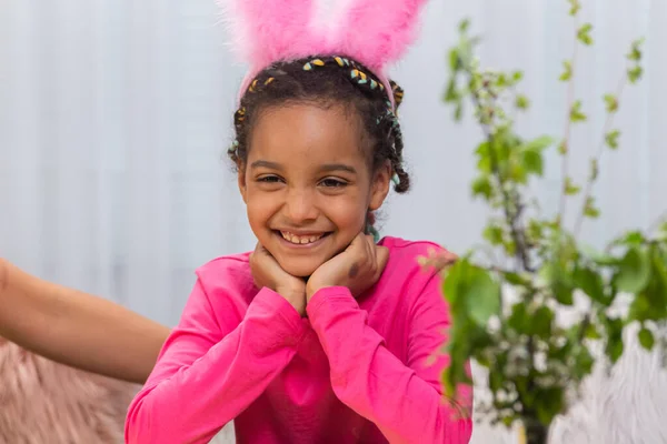 Una bambina carina in divertenti orecchie di coniglio rosa sorride. Bambino afroamericano in posa sullo sfondo di una stanza luminosa. Buona Pasqua. Da vicino.. — Foto Stock