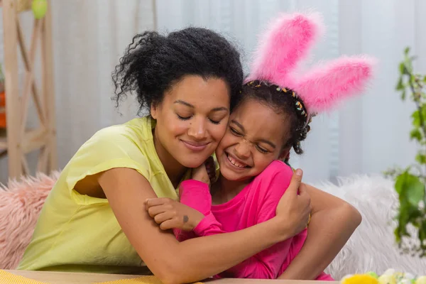 Mamma e figlia con divertenti orecchie da coniglio si abbracciano e sorridono felicemente. Donna afroamericana e bambina sono seduti a tavola in una stanza decorata a festa a casa. Da vicino.. — Foto Stock