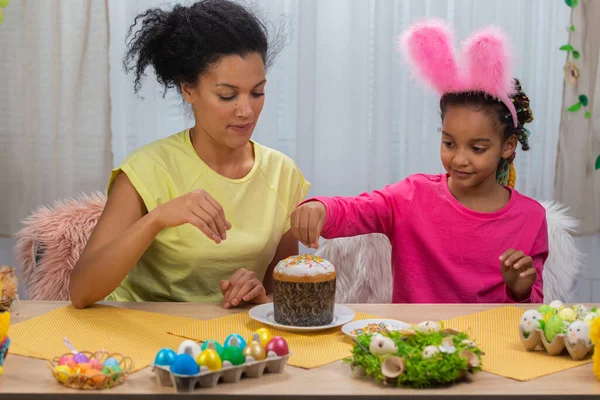 Mamma e figlia con divertenti orecchie di coniglio decorano la torta di Pasqua con dolci caramelle. La donna afroamericana e la bambina sono sedute a tavola in una stanza decorata a festa. Buona Pasqua. Da vicino.. — Foto Stock