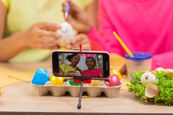 Mamá y su hija con orejas de conejito divertido filmando en el teléfono que pintan huevos. Mujer afroamericana y niña están sentadas a la mesa en una habitación festivamente decorada. Feliz Pascua. De cerca.. —  Fotos de Stock