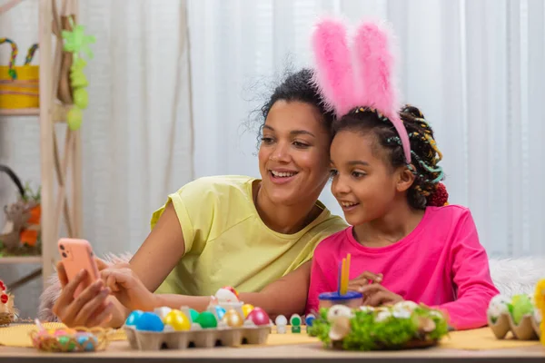 Mamma e figlia con le orecchie divertenti coniglietto parlare in una videochiamata utilizzando il telefono. La donna afroamericana e la bambina sono sedute a tavola in una stanza decorata a festa. Buona Pasqua. Da vicino.. — Foto Stock