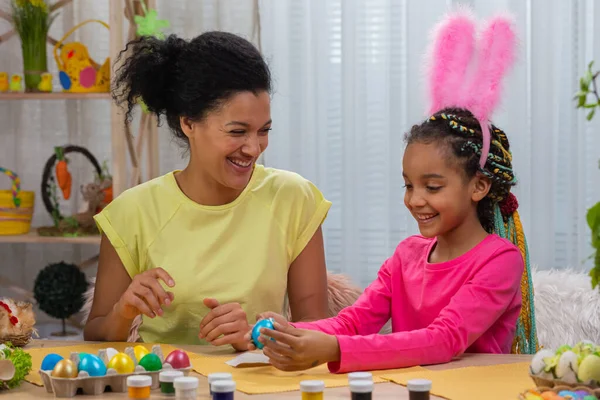 Mor og datter med rare kaninører som limer bilder på fargede egg. Afroamerikansk kvinne og liten jente sitter ved bordet i et dekorert rom hjemme. Gledelig østlig. Lukk. royaltyfrie gratis stockbilder