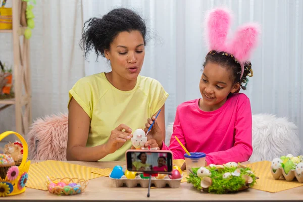 Mor og datter med rare kaninører som filmer på telefonen og maler egg. Afroamerikansk kvinne og liten jente sitter ved bordet i et festlig dekorert rom. Gledelig østlig. Lukk. stockbilde