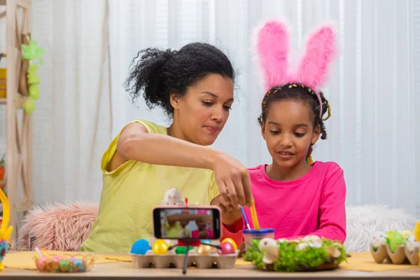 Mor og datter med rare kaninører som filmer på telefonen og maler egg. Afroamerikansk kvinne og liten jente sitter ved bordet i et festlig dekorert rom. Gledelig østlig. Lukk. stockfoto