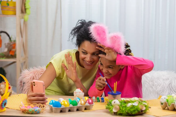 Mamma e figlia con le orecchie divertenti coniglietto parlare in una videochiamata utilizzando il telefono. La donna afroamericana e la bambina sono sedute a tavola in una stanza decorata a festa. Buona Pasqua. Da vicino.. — Foto Stock