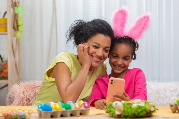 Mamma e figlia con divertenti orecchie da coniglio si scattano selfie e si divertono. Donna afroamericana e bambina sono seduti a tavola in una stanza decorata a festa a casa. Buona Pasqua. Da vicino.. — Foto Stock