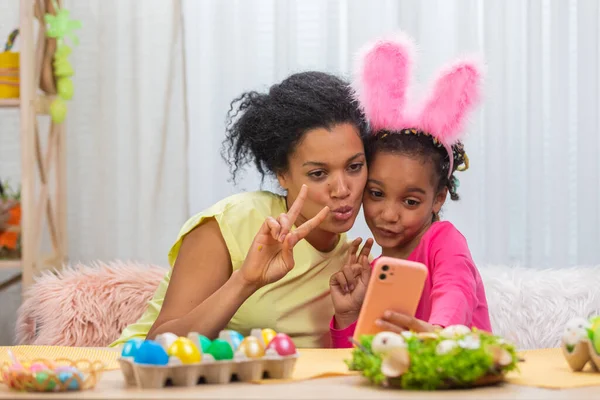 Mamma e figlia con divertenti orecchie da coniglio si scattano selfie e si divertono. Donna afroamericana e bambina sono seduti a tavola in una stanza decorata a festa a casa. Buona Pasqua. Da vicino.. — Foto Stock